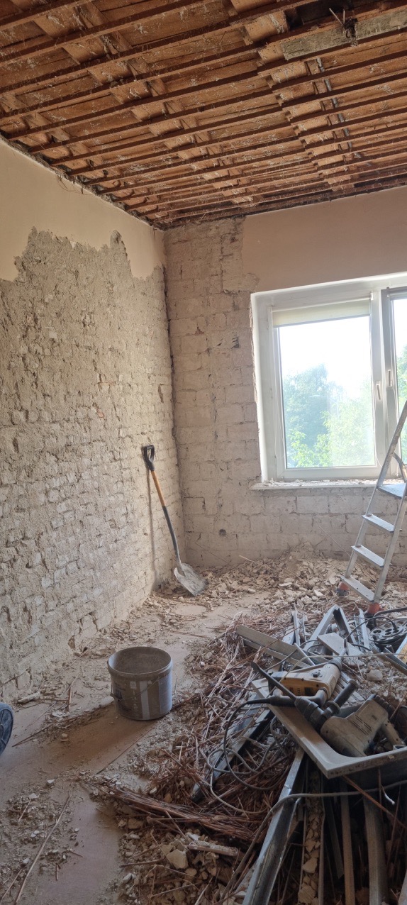 Pokój w trakcie remontu: otynkowana ściana z cegły, drewniany sufit, gruz na podłodze, drabina oparta o ścianę przy oknie, wiadro i łopata w rogu.