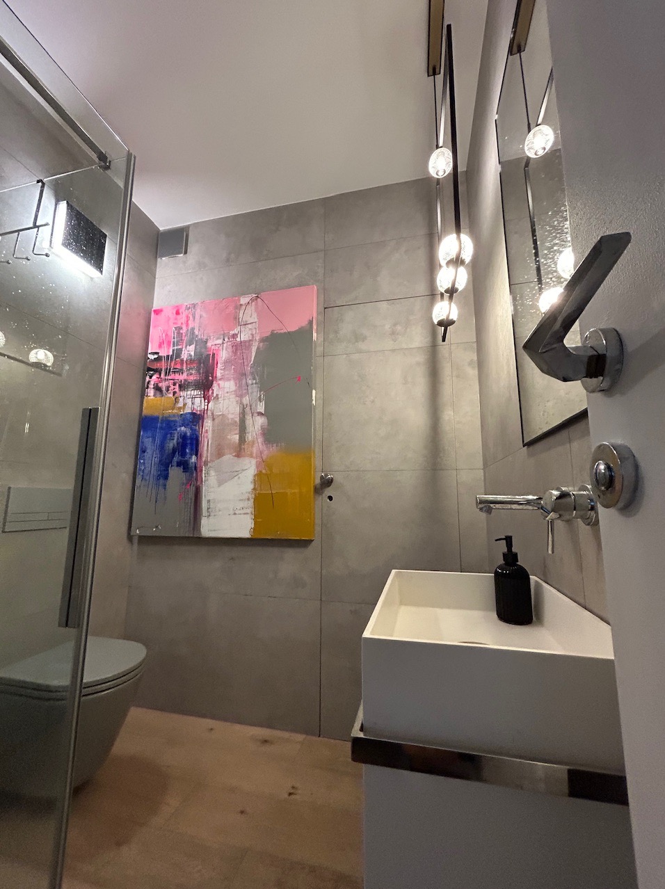 Nowoczesna łazienka z szarą płytką, wiszącą toaletą za szklaną kabiną, białą umywalką nablatową, abstrakcyjnym obrazem na ścianie i designerską lampą wiszącą z kulistymi kloszami.