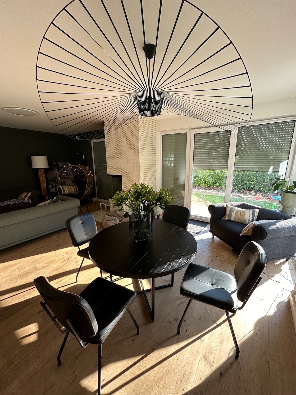 Nowoczesne wnętrze domu z okrągłym, czarnym stołem jadalnym, czarnymi krzesłami i designerską lampą sufitową w kształcie wachlarza, oświetlone promieniami słońca padającymi przez okno.