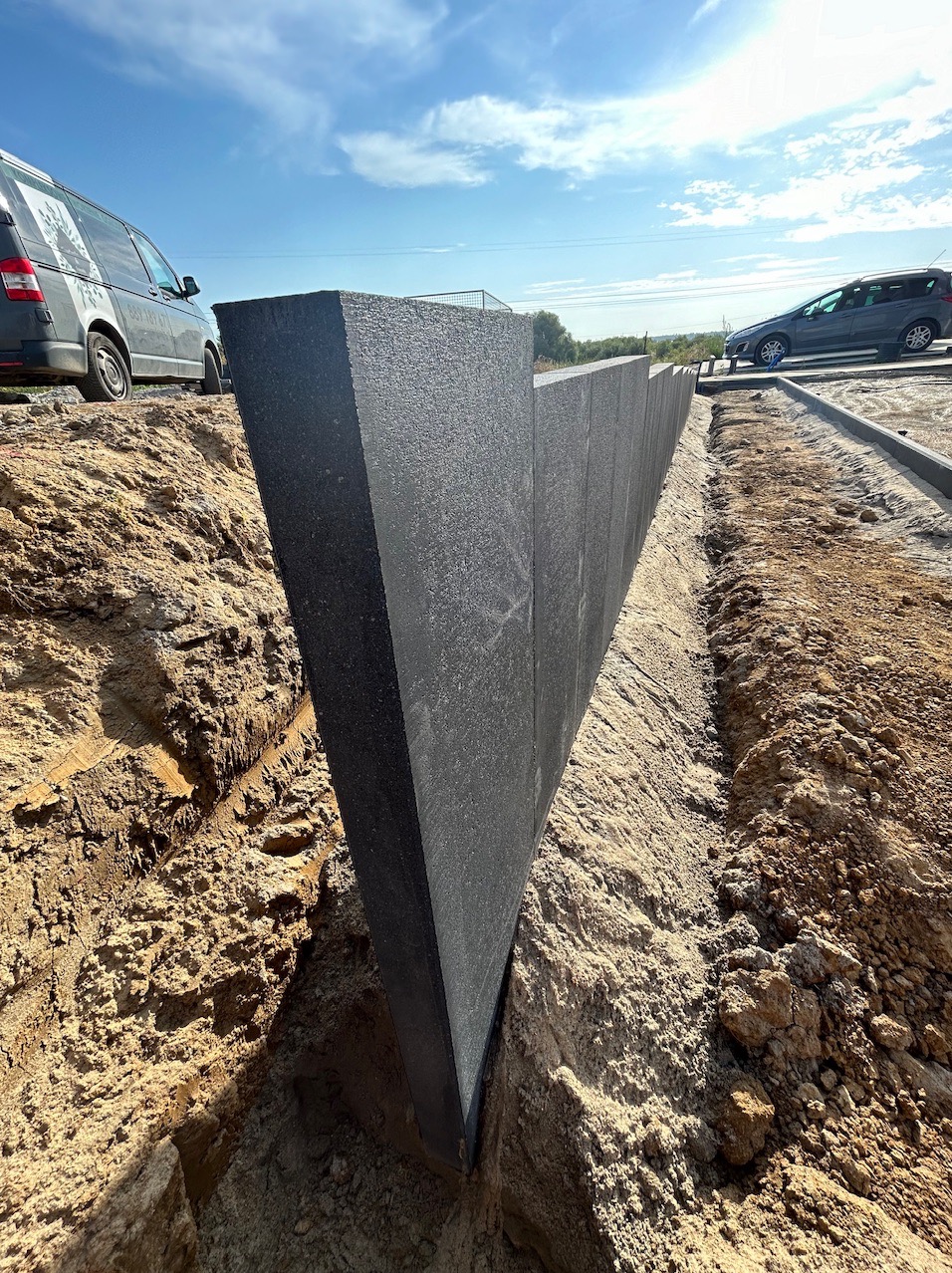 Czarne, trójkątne panele z tworzywa sztucznego wkopane w ziemię, tworzące ogrodzenie lub zabezpieczenie terenu budowy, widoczne samochody w tle.