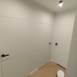 PATRYK ŻYWICA WIZJA REMONT - Nowoczesne, białe drzwi wewnętrzne z czarnymi klamkami w minimalistycznym przedpokoju z podłogą w jodełkę i wiaderkiem farby. Oświetlenie sufitowe.