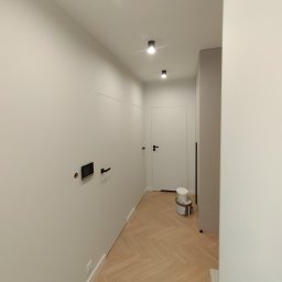 PATRYK ŻYWICA WIZJA REMONT - Korytarz w jasnych barwach po remoncie: parkiet w jodełkę, białe ściany z czarnymi detalami (gniazdko, uchwyt), drzwi wewnętrzne. Widoczne puszki z farbą.