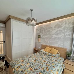 PATRYK ŻYWICA WIZJA REMONT - Jasna sypialnia z tapetą w drzewa, białymi drzwiami z szybami i szafą w zabudowie. Pies rasy owczarek australijski siedzi na podłodze przy drzwiach.