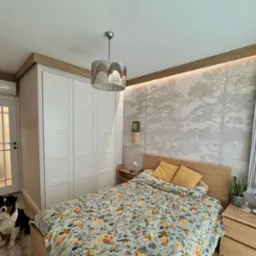 Jasna sypialnia z tapetą w drzewa, białymi drzwiami z szybami i szafą w zabudowie. Pies rasy owczarek australijski siedzi na podłodze przy drzwiach.