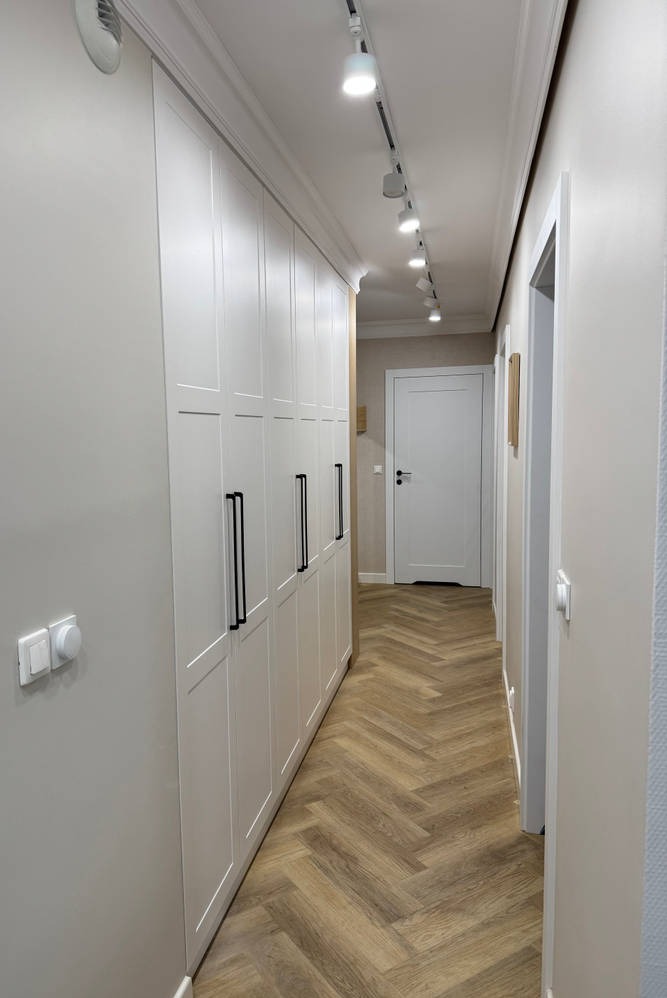 Korytarz z zabudową szaf w kolorze białym i parkietem ułożonym we wzór jodełki. Oświetlenie sufitowe punktowe. Widok na drzwi na końcu korytarza.