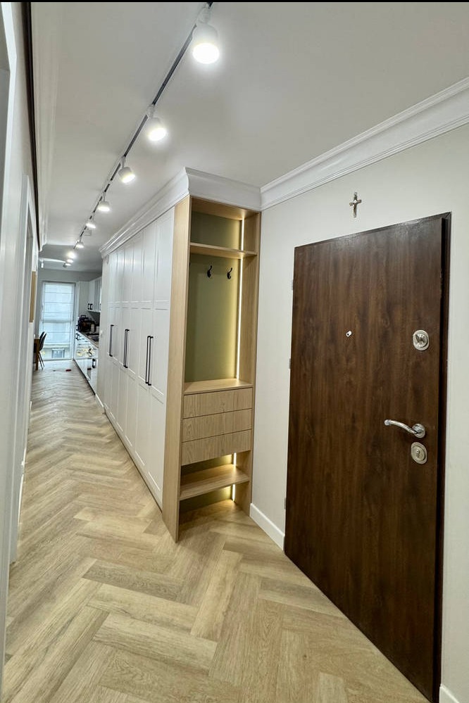 Nowoczesny przedpokój z szafą wnękową z oświetleniem LED, drewnianymi panelami na podłodze ułożonymi w jodełkę i ciemnymi drzwiami wejściowymi z metalowym wizjerem.