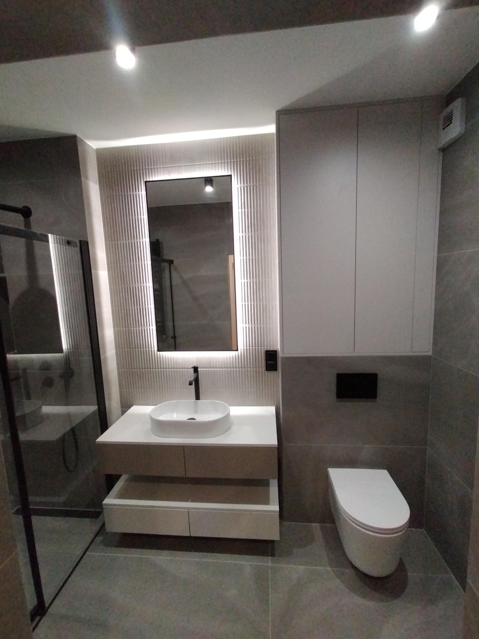 Nowoczesna łazienka w szarościach z podświetlanym lustrem, umywalką nablatową i wiszącą toaletą. Minimalistyczne szafki i kabina prysznicowa.