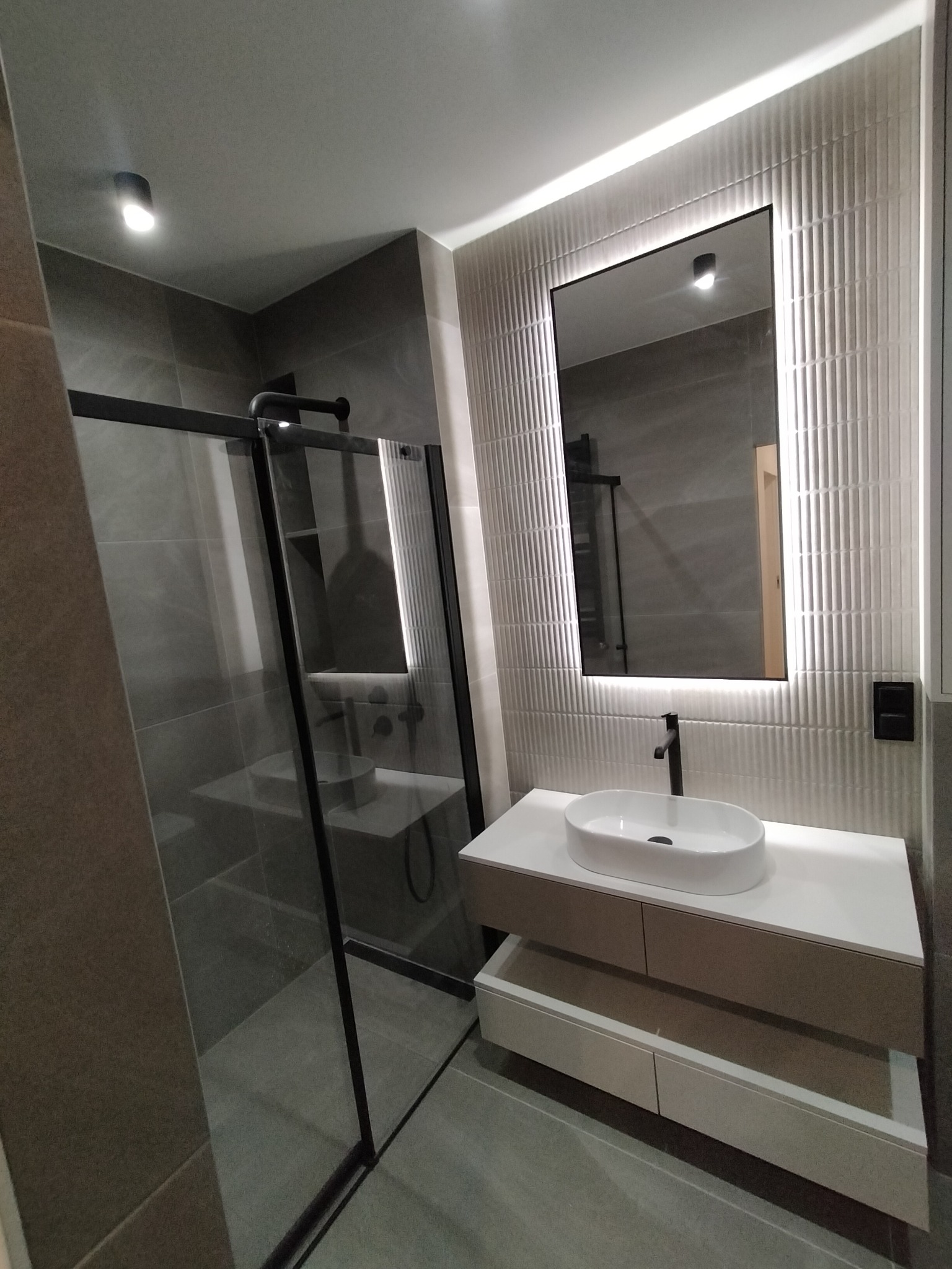 Nowoczesna łazienka po remoncie: kabina prysznicowa typu walk-in z czarnymi ramami, umywalka nablatowa na szafce i podświetlane lustro na ścianie z dekoracyjną glazurą.