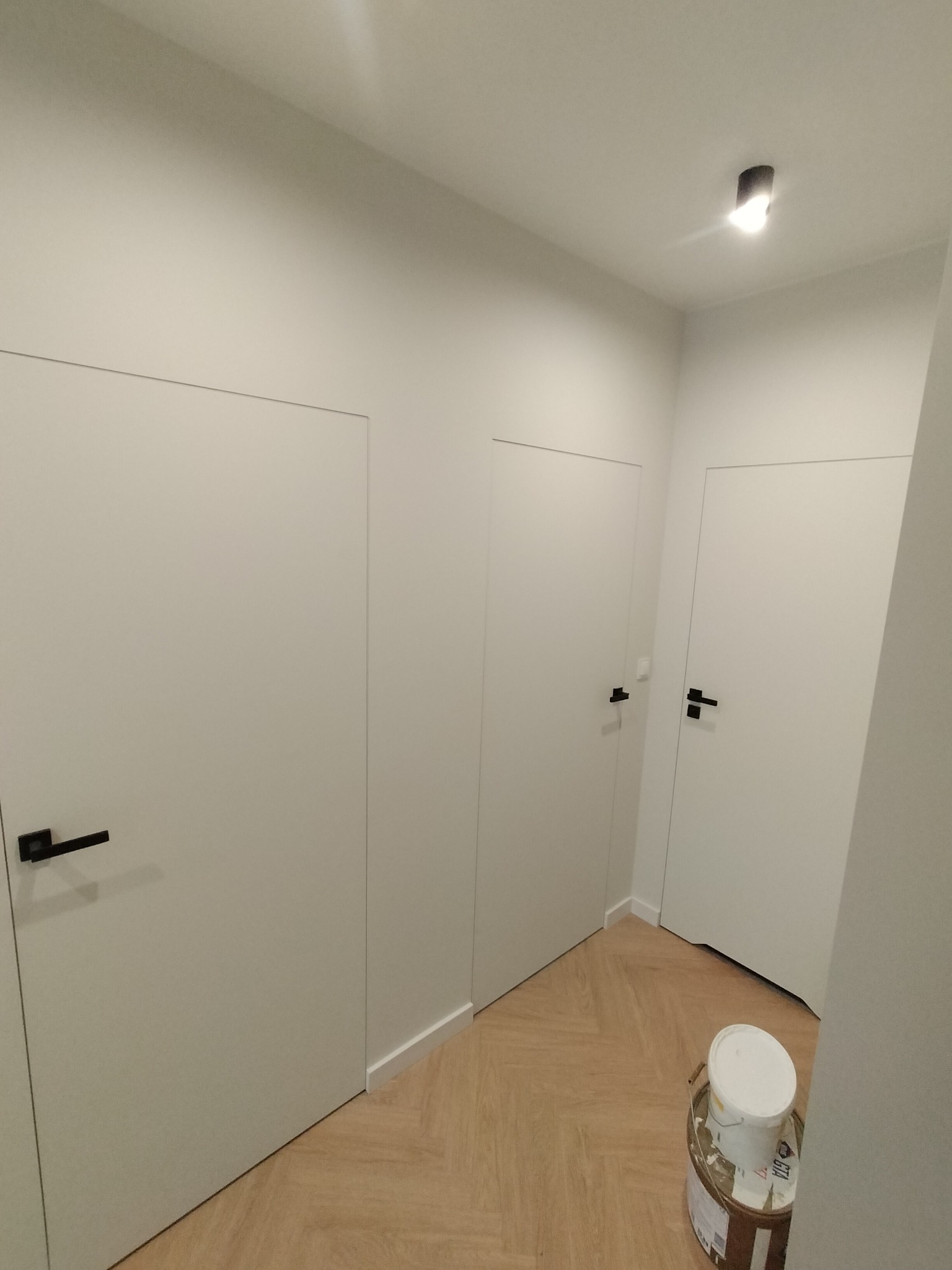 Nowoczesne, białe drzwi wewnętrzne z czarnymi klamkami w minimalistycznym przedpokoju z podłogą w jodełkę i wiaderkiem farby. Oświetlenie sufitowe.
