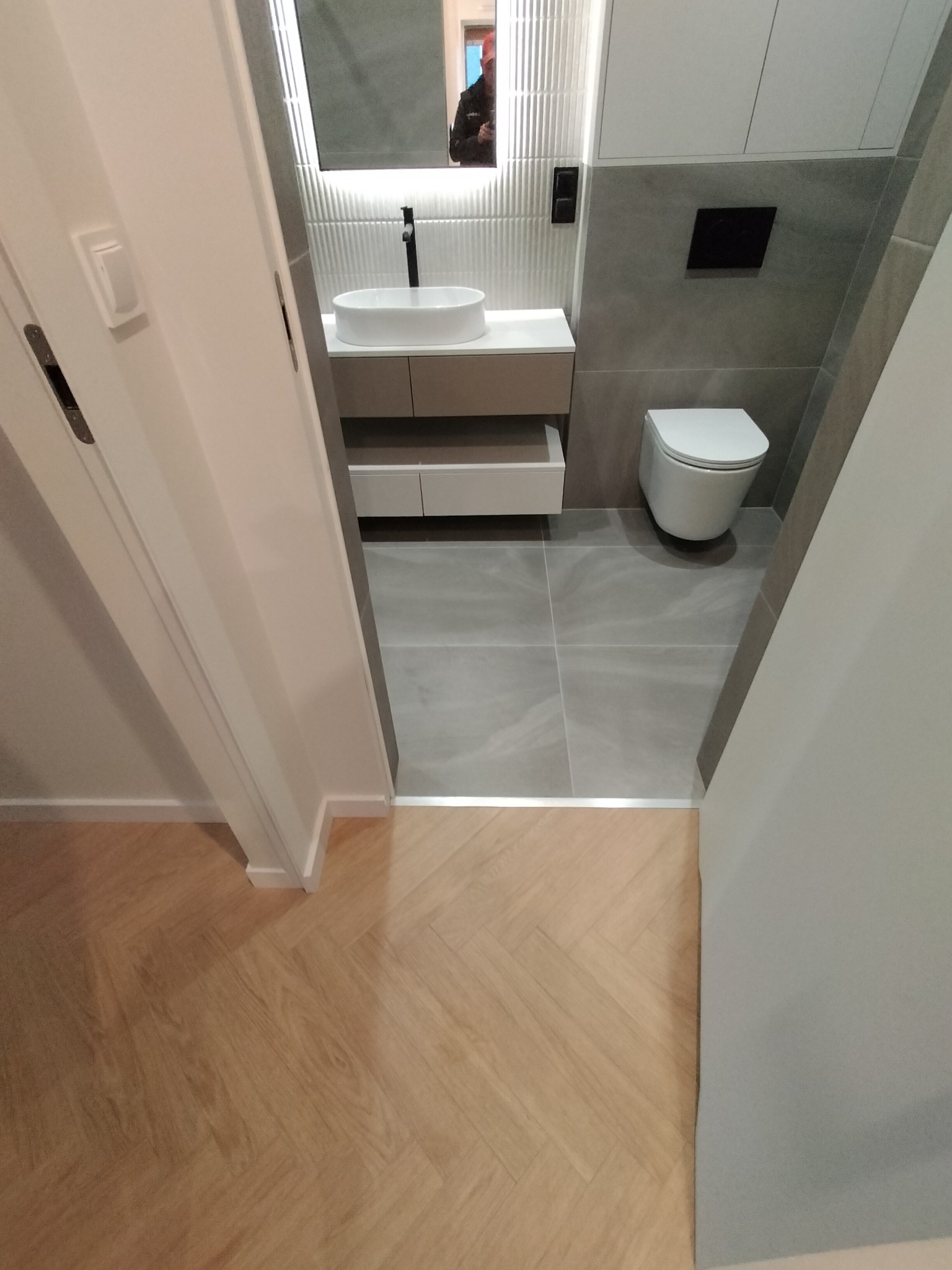 Nowoczesna łazienka z umywalką nablatową, podwieszaną toaletą i szarą glazurą, widziana zza uchylonych drzwi. Parkiet w jodełkę poza łazienką.