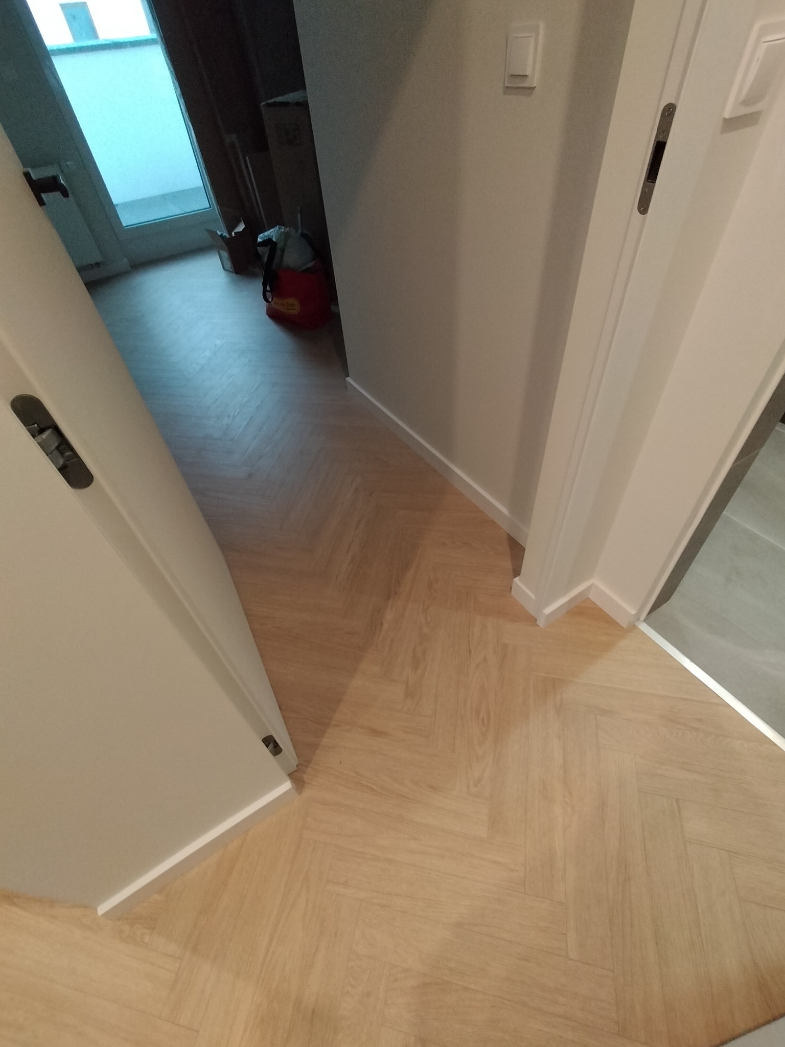 Ukończone układanie jasnych paneli podłogowych w jodełkę francuską, widoczne fragmenty białych drzwi i listew przypodłogowych. Estetyczne wykończenie.