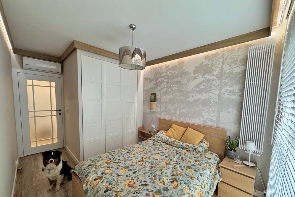 Jasna sypialnia z tapetą w drzewa, białymi drzwiami z szybami i szafą w zabudowie. Pies rasy owczarek australijski siedzi na podłodze przy drzwiach.