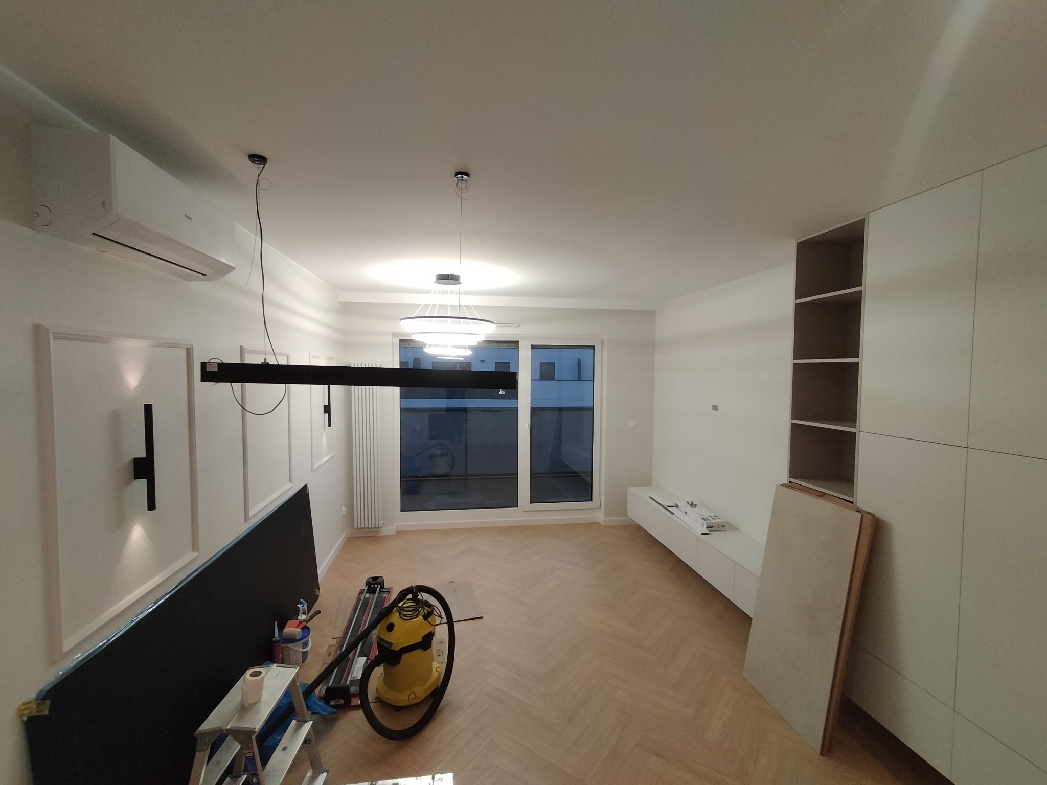 Jasne wnętrze w trakcie wykończenia: parkiet w jodełkę, białe ściany z dekoracyjnymi listwami, częściowo zamontowane meble i narzędzia pracy. Widok na okno balkonowe.