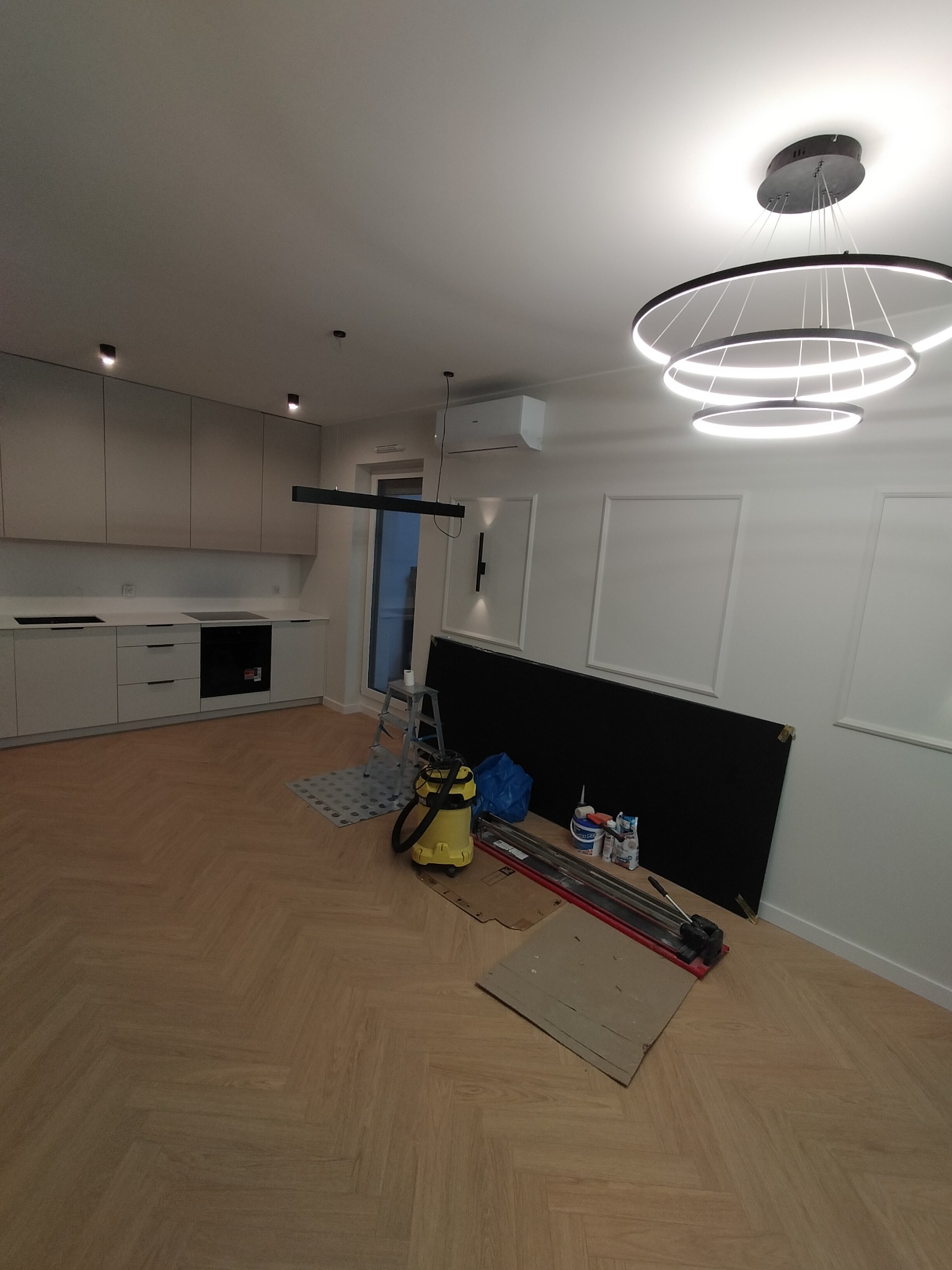 Jasne panele ułożone w jodełkę francuską w przestronnym wnętrzu z kuchnią. Widoczne narzędzia glazurnicze i drabina. Nowoczesna lampa sufitowa.