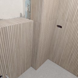 Szymon Witt - Montaż dekoracyjnych paneli ściennych imitujących drewno w łazience, widoczne gniazdka elektryczne i otwory na instalacje, fragment surowej ściany z zaprawą.