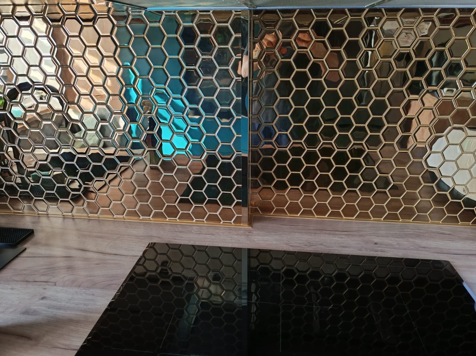Dwie panele dekoracyjne z heksagonalnymi wzorami w odcieniach złota i miedzi, jeden panel odbija niebieskie światło, ułożone nad blatem i płytą indukcyjną z podobnym wzorem.