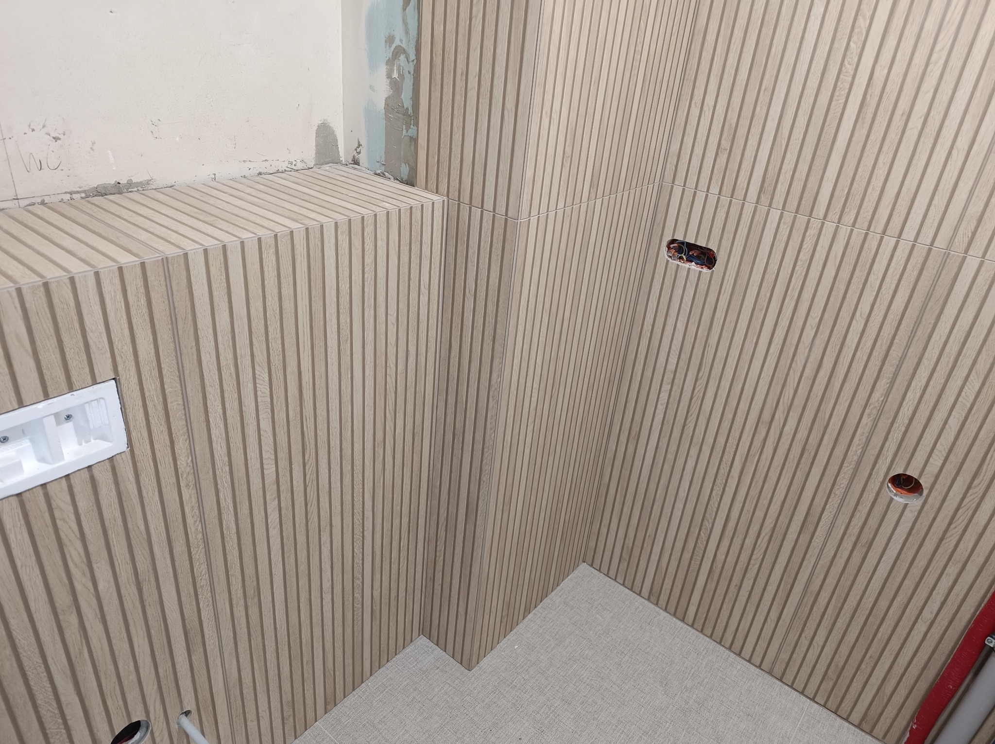 Montaż dekoracyjnych paneli ściennych imitujących drewno w łazience, widoczne gniazdka elektryczne i otwory na instalacje, fragment surowej ściany z zaprawą.