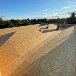 Wymiana dachu z płyt OSB w Mogilnie Dużym, widok z g&oacute;ry na konstrukcję przed położeniem pokrycia, komin z cegły klinkierowej w tle, słoneczny dzień.