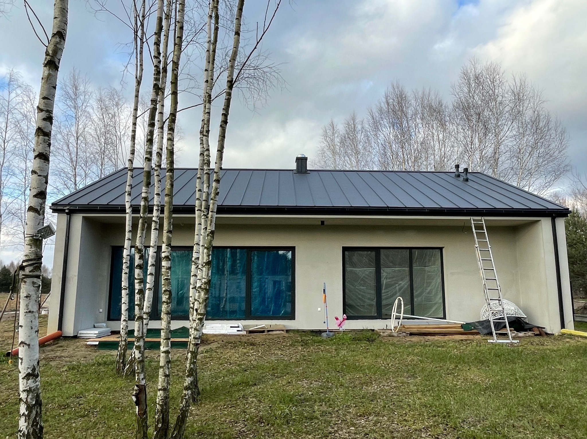 Nowoczesny dom z ciemnoszarym dachem blaszanym na rąbek stojący, drabina oparta o ścianę, brzozy na pierwszym planie