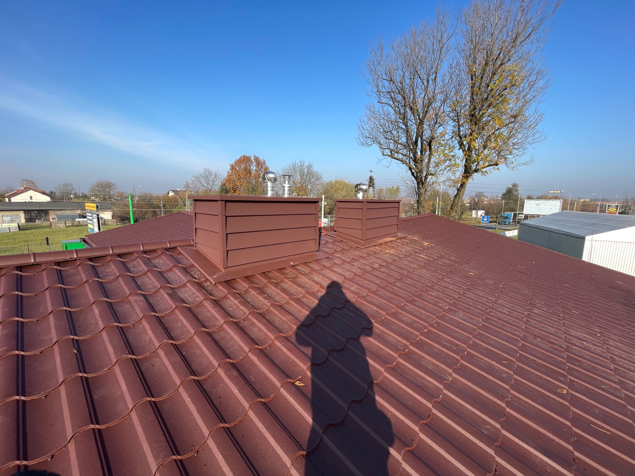 Ciemnoczerwony dach z blachodachówki z dwoma kominami w kształcie pudełek, cień osoby fotografującej na dachu, w tle drzewa i błękitne niebo.