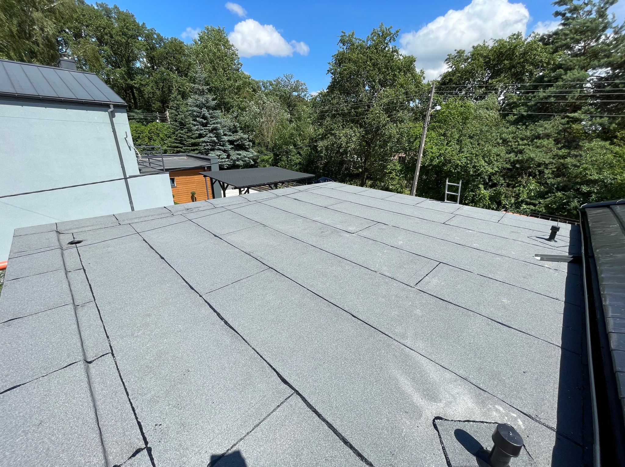 Szary dach pokryty papą termozgrzewalną na tle zielonych drzew i błękitnego nieba z chmurami. Widoczna drabina oparta o drzewo w oddali.