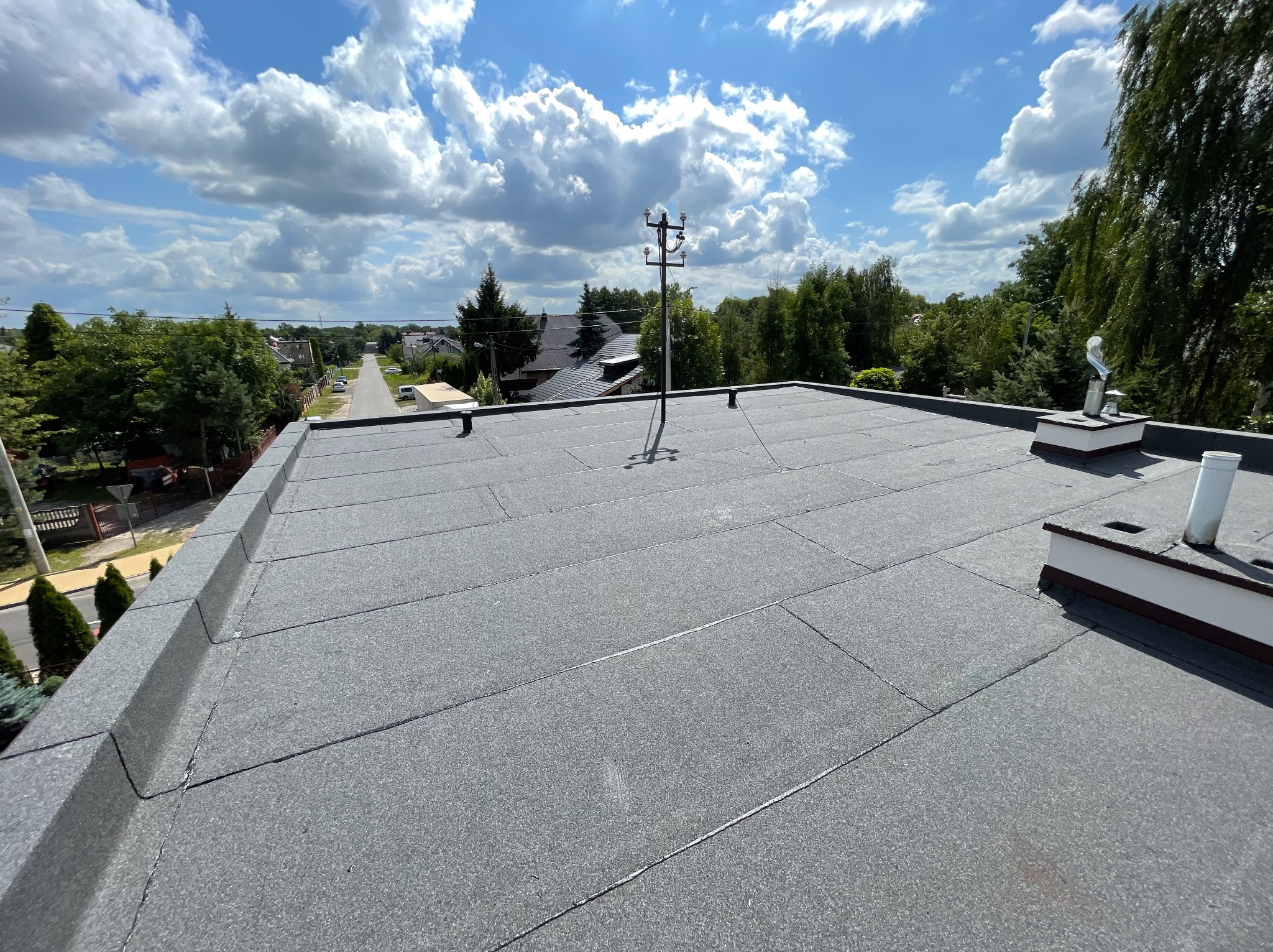 Widok z góry na płaski dach pokryty papą termozgrzewalną z widocznymi kominami wentylacyjnymi i słupem elektrycznym na tle błękitnego nieba z obłokami.