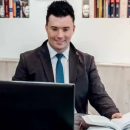 Uśmiechnięty mężczyzna w garniturze i krawacie siedzi przy biurku z laptopem i otwartą książką, półki z książkami i dekoracjami w tle.