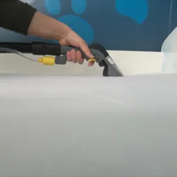 Czyszczenie materaca za pomocą odkurzacza piorącego z dyszą, widoczna ręka operatora i fragment ściany z dekoracyjnym malunkiem.