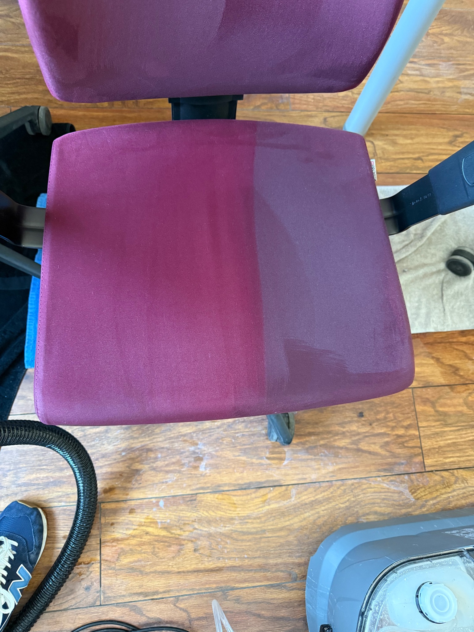 Krzesło biurowe z bordową tapicerką, widoczna różnica w czystości połowy siedziska po praniu ekstrakcyjnym, obok odkurzacz piorący i wąż ssący na drewnianej podłodze.