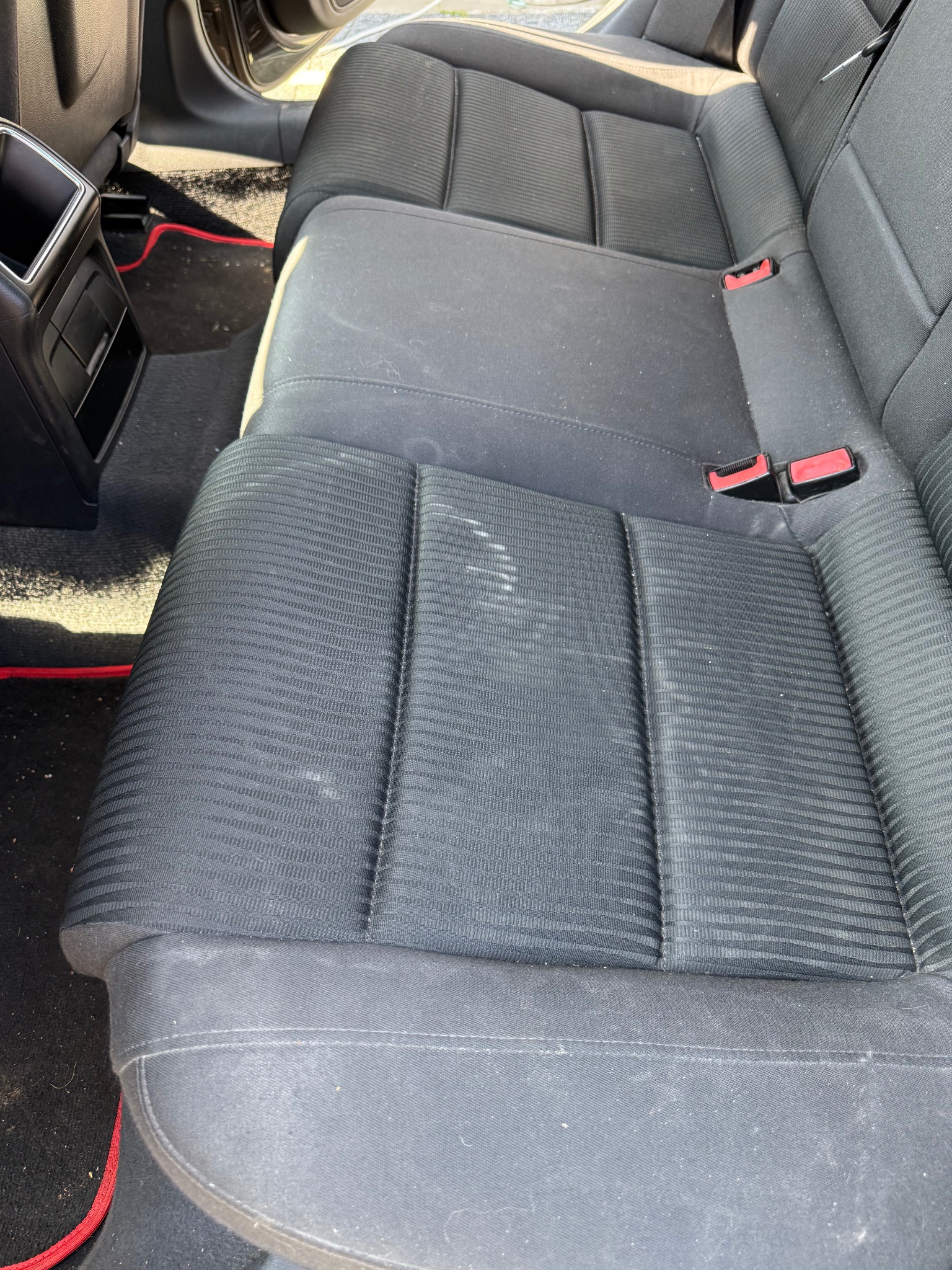 Brudna, tekstylna tylna kanapa samochodowa z widocznymi plamami i okruchami, czarny dywanik z czerwonym obszyciem na podłodze, uchwyt na napoje w tle.
