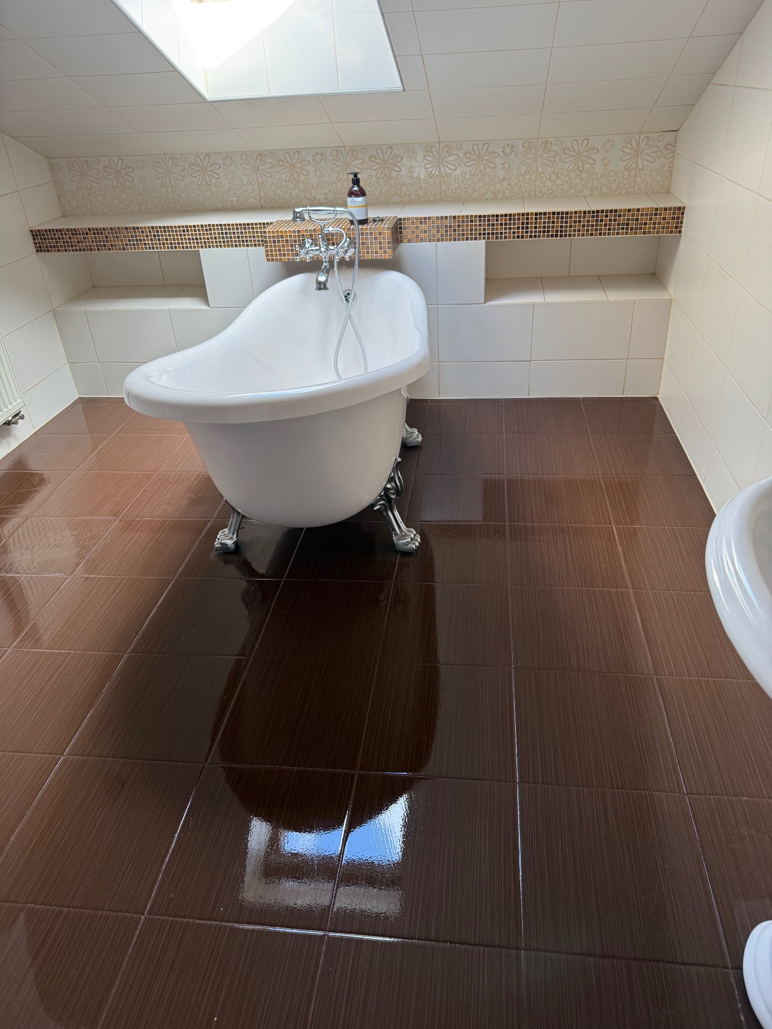 Błyszcząca, brązowa podłoga w łazience w Święciechowie z białą wanną na lwich nóżkach i dekoracyjną półką nad wanną.