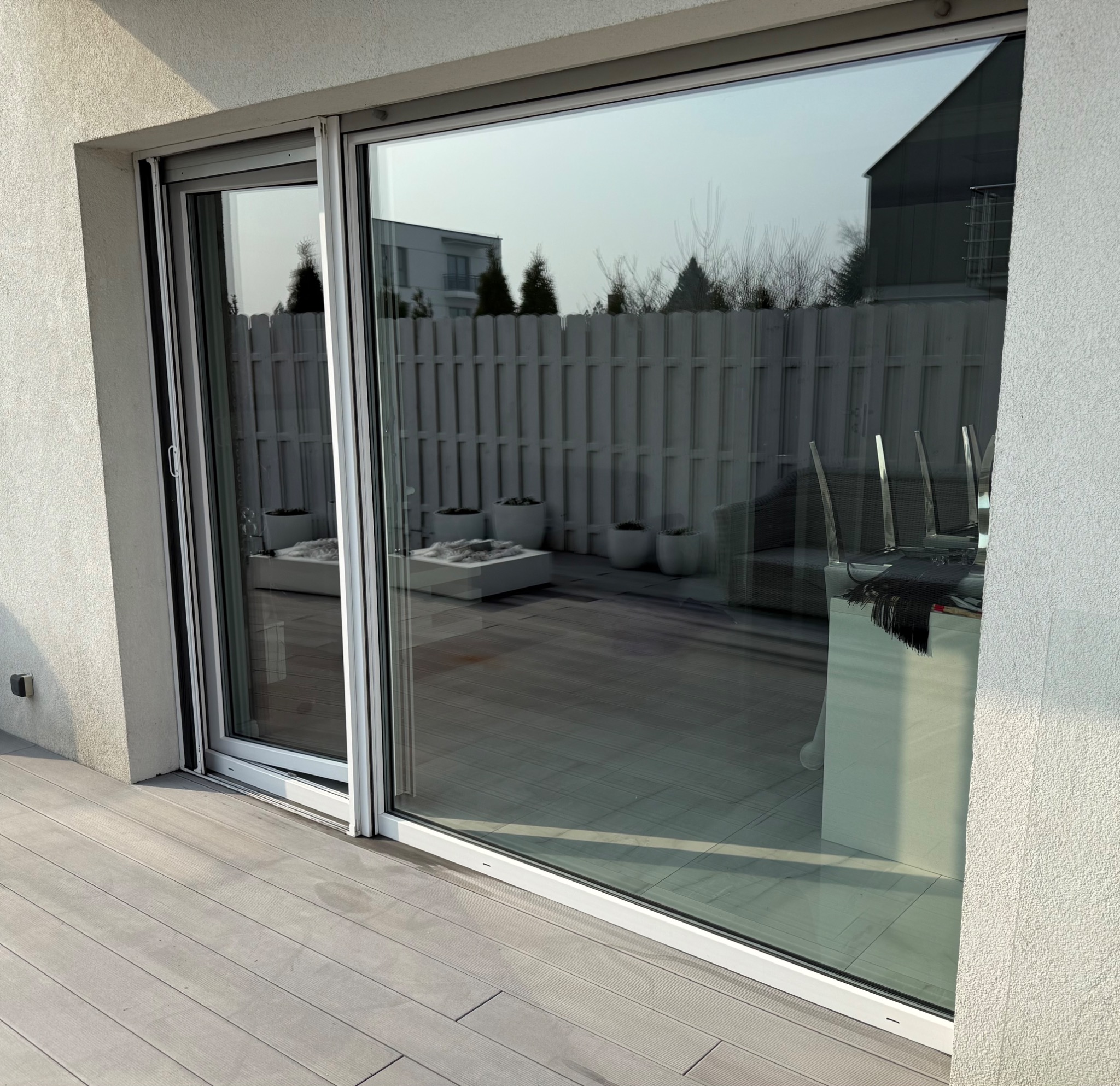 Przesuwne okno balkonowe z widokiem na taras z drewnianą podłogą, białym ogrodzeniem i dekoracyjnymi donicami z roślinami.