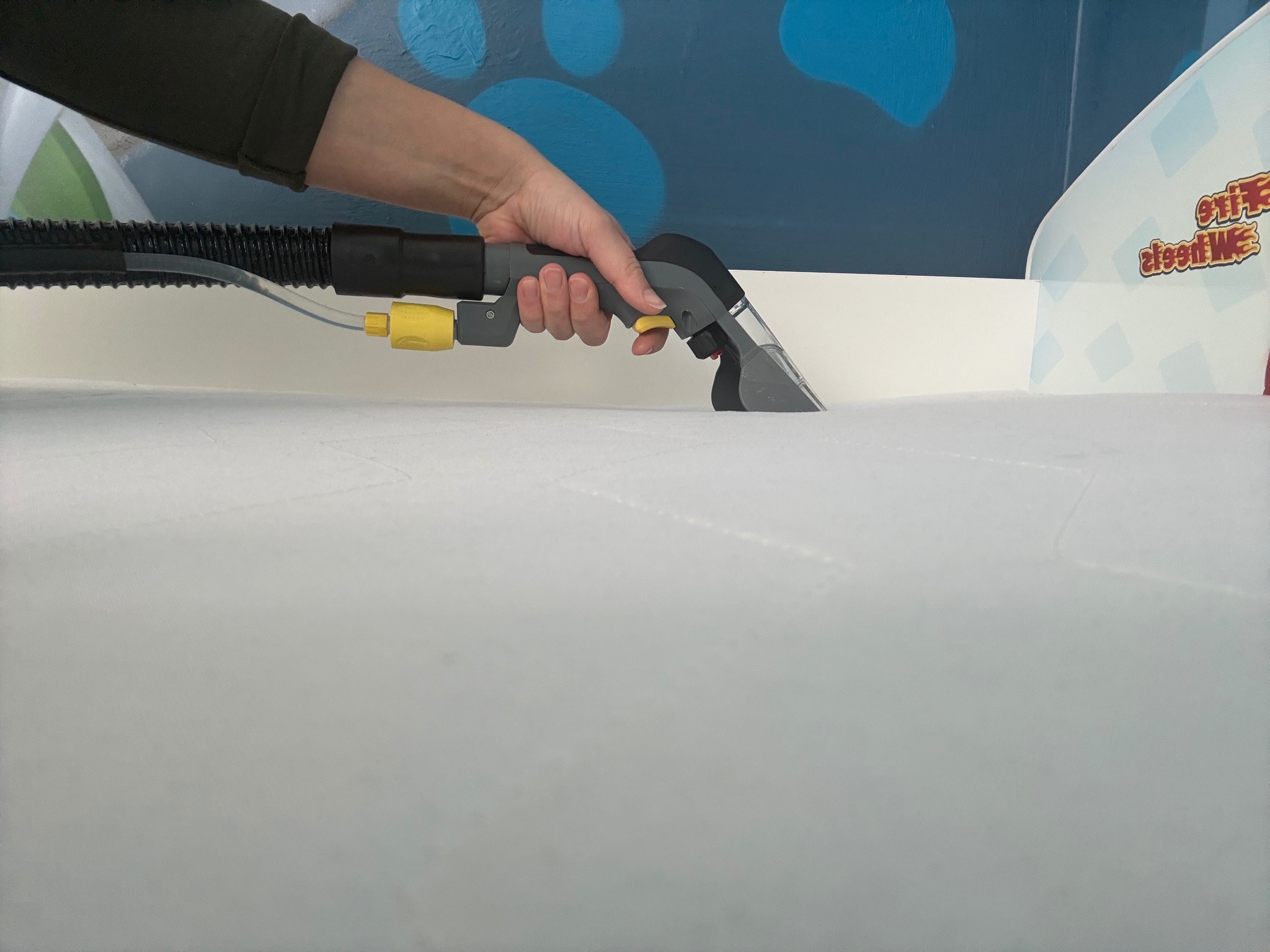 Czyszczenie materaca za pomocą odkurzacza piorącego z dyszą, widoczna ręka operatora i fragment ściany z dekoracyjnym malunkiem.