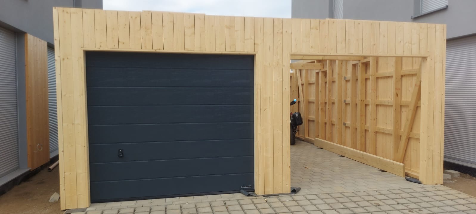 Konstrukcja garażu z drewna w trakcie budowy, widoczna brama garażowa segmentowa w kolorze antracytowym i szkielet z desek po prawej stronie, na zewnątrz budynku.