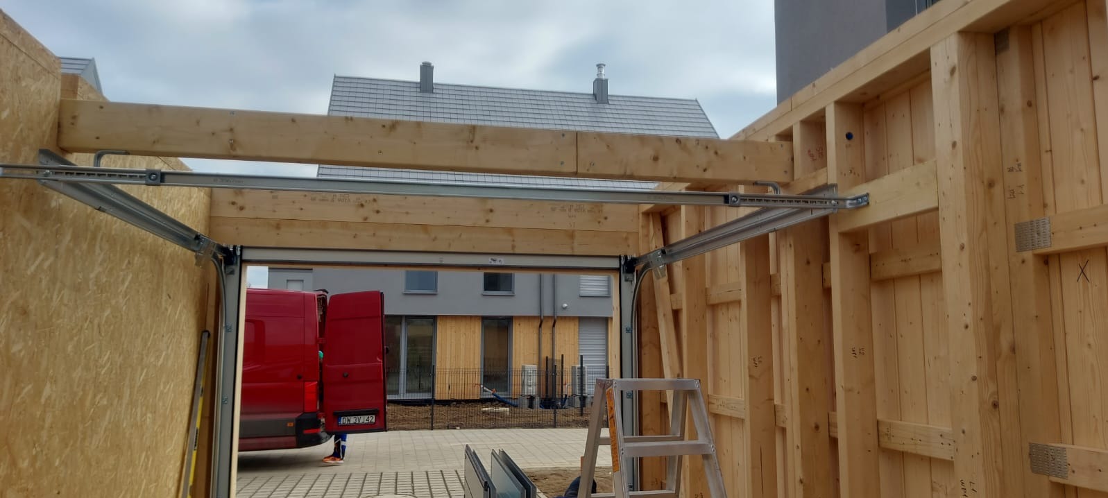 Wnętrze garażu w surowym stanie domu szkieletowego, widoczne drewniane belki konstrukcyjne, prowadnice bramy garażowej i czerwony samochód dostawczy w tle.