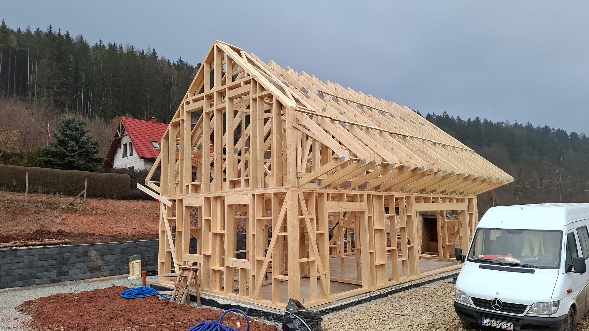 Konstrukcja domu w systemie szkieletowym w trakcie budowy, widoczne drewniane elementy nośne ścian i dachu, na zewnątrz zaparkowany biały bus, w tle las i dom z czerwonym dachem.