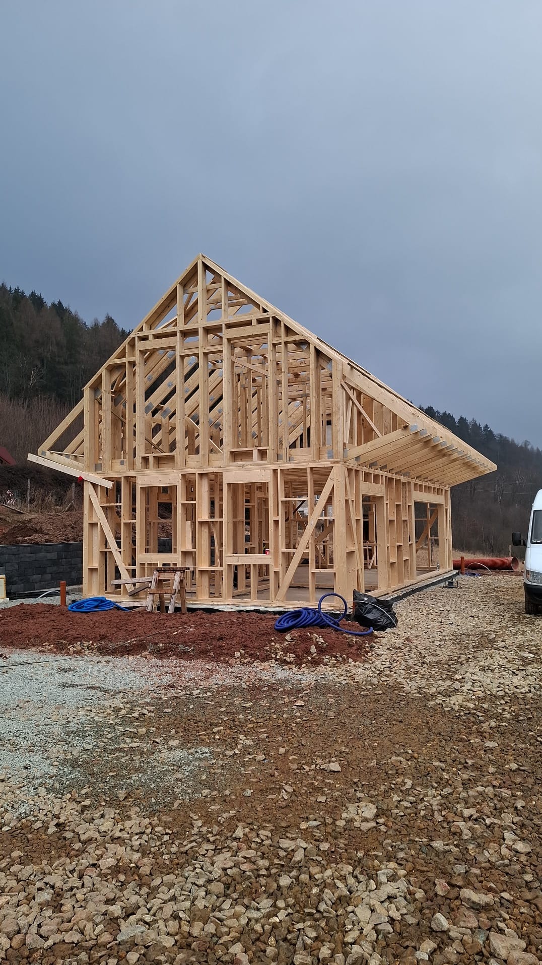 Konstrukcja drewniana domu w trakcie budowy, widoczne ściany i dach, w tle las i pochmurne niebo.