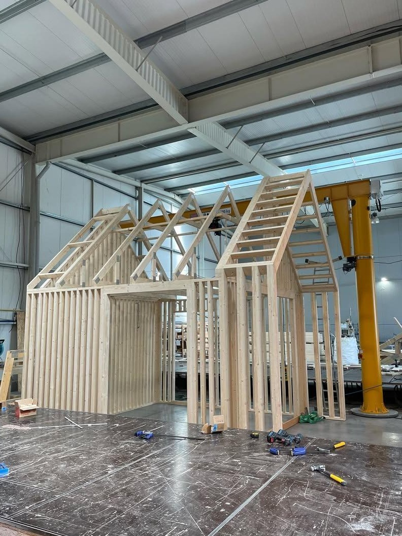 Konstrukcja szkieletowa domu w hali produkcyjnej, widoczny żółty suwnicowy dźwig, narzędzia na podłodze, jasne drewno.