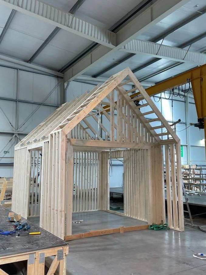 Konstrukcja szkieletowa domu w trakcie montażu w hali produkcyjnej, widoczne narzędzia i elementy drewniane, żółty dźwig w tle.