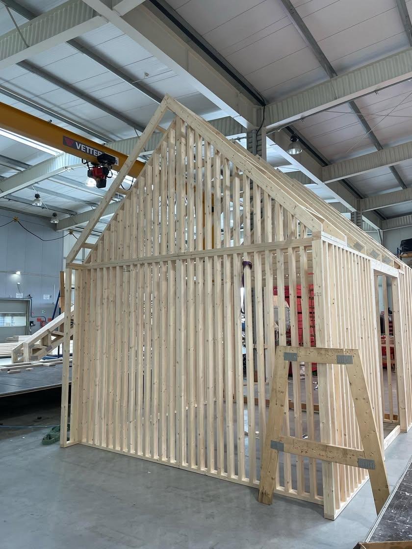 Surowa konstrukcja domu z drewnianych belek, widoczna w hali produkcyjnej z suwnicą pod sufitem.