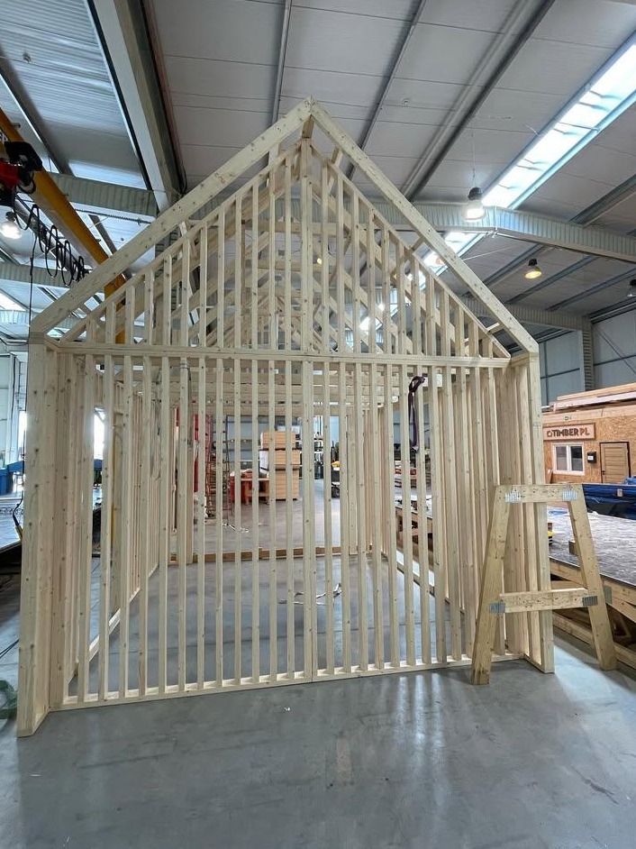 Wnętrze hali produkcyjnej z widoczną konstrukcją szkieletową domu, drewniane elementy połączone, w tle widoczny częściowo gotowy budynek z logo firmy.