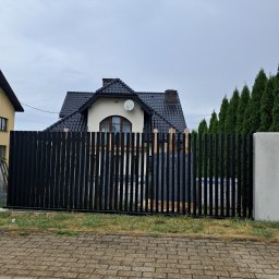 Tomasz Nabagło - Częściowo zamontowane, czarne, pionowe ogrodzenie metalowe na tle domu z ciemnym dachem i tujami. Widoczne drewniane elementy montażowe.