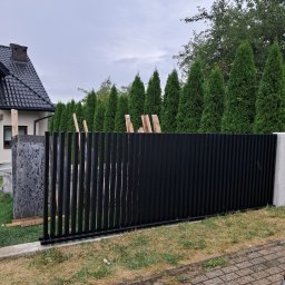 Tomasz Nabagło - Częściowo zamontowane, czarne, pionowe panele ogrodzenia metalowego na tle zielonych drzew i domu z ciemnym dachem. Widoczne elementy montażowe.