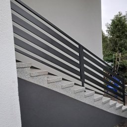 Tomasz Nabagło - Zewnętrzne schody z szarego kamienia z nowoczesną, poziomą balustradą z czarnych, metalowych profili. Minimalistyczny design, jasne tło budynku.