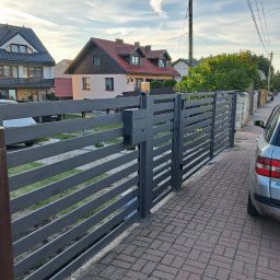 Tomasz Nabagło - Nowoczesne, poziome ogrodzenie metalowe w kolorze grafitowym, zintegrowane ze skrzynką na listy. Widok z zewnątrz posesji, kostka brukowa, domy w tle.