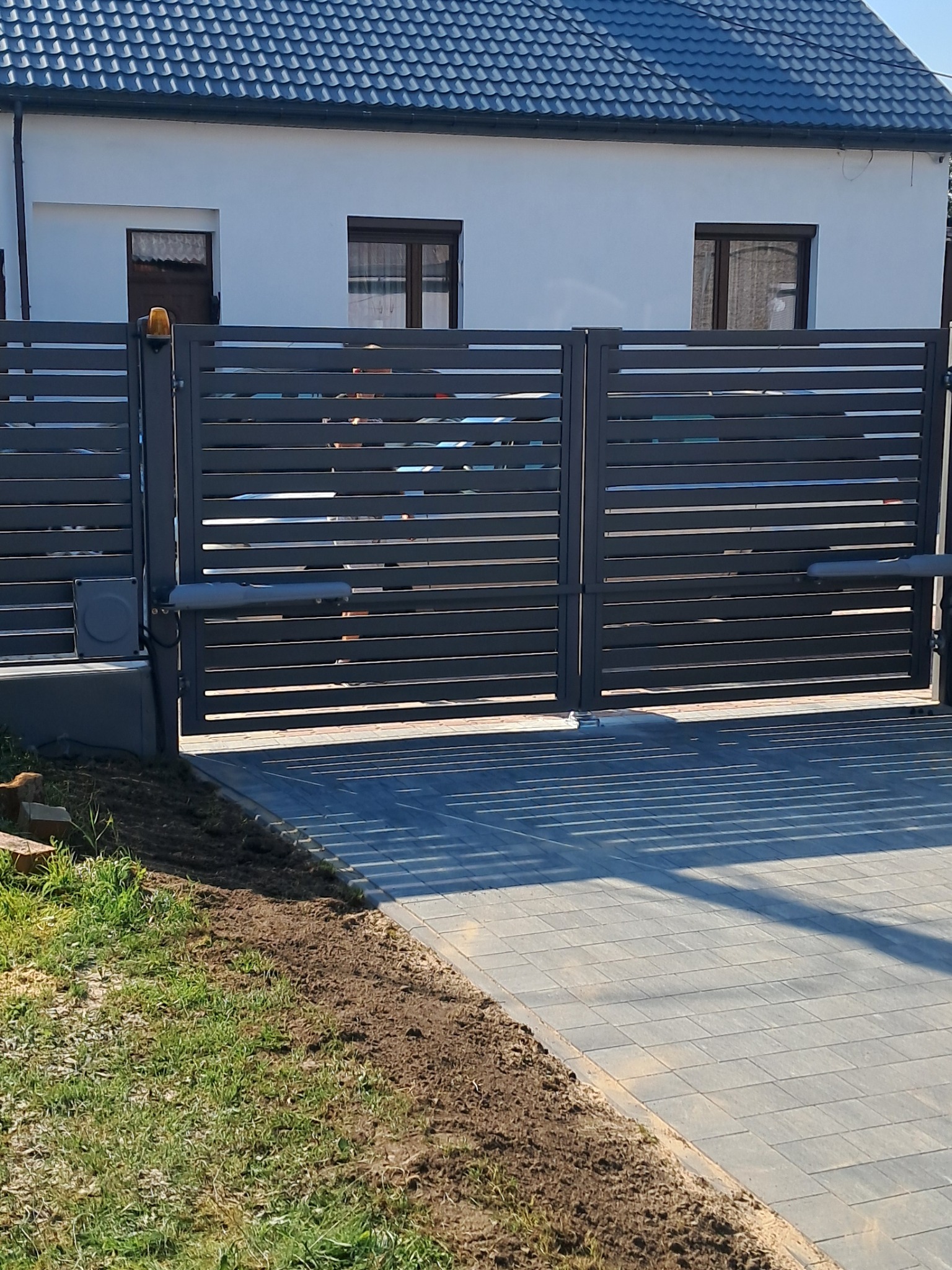 Nowoczesna, przesuwna brama metalowa w kolorze grafitowym z automatyką, zamontowana przed domem z ciemną dachówką. Widoczne poziome panele i kostka brukowa.