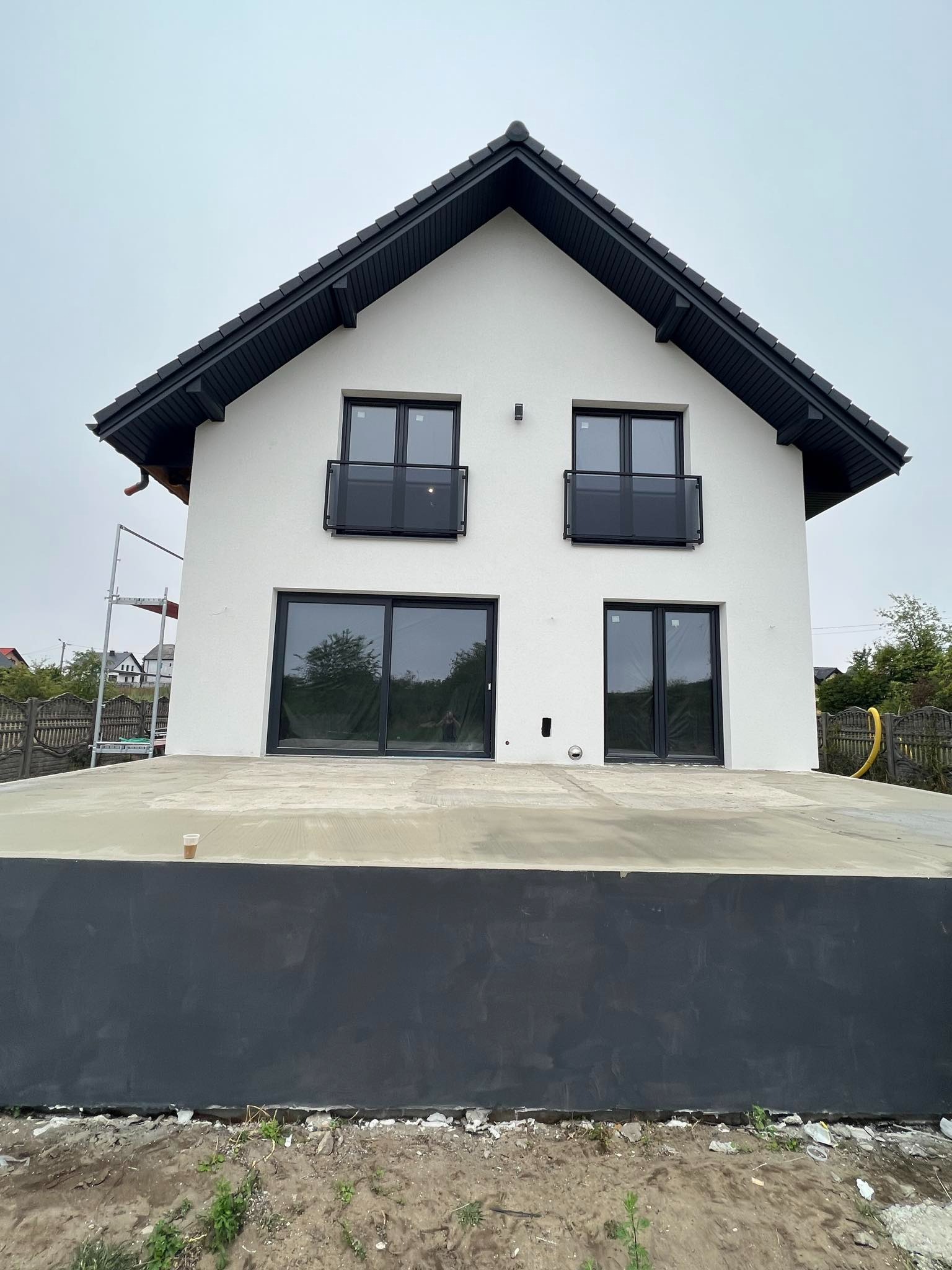 Nowoczesny dom z białymi ścianami i czarnym dachem. Widoczne czarne balustrady balkonowe i duże okna. Na pierwszym planie betonowa podstawa budynku.