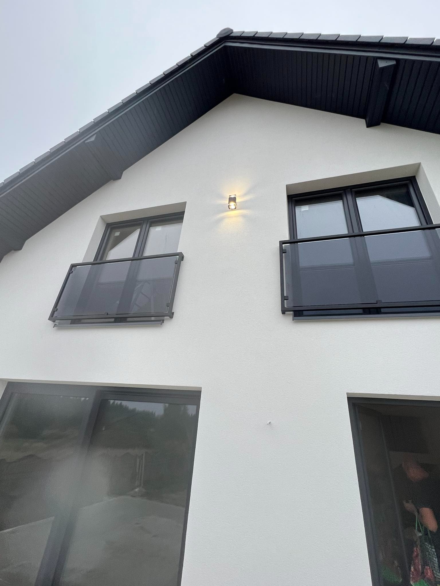 Biała fasada domu z dwoma balkonami z balustradami szklanymi i czarnymi ramami okiennymi. Nad oknami czarne wykończenie dachu. Ujęcie z dołu.