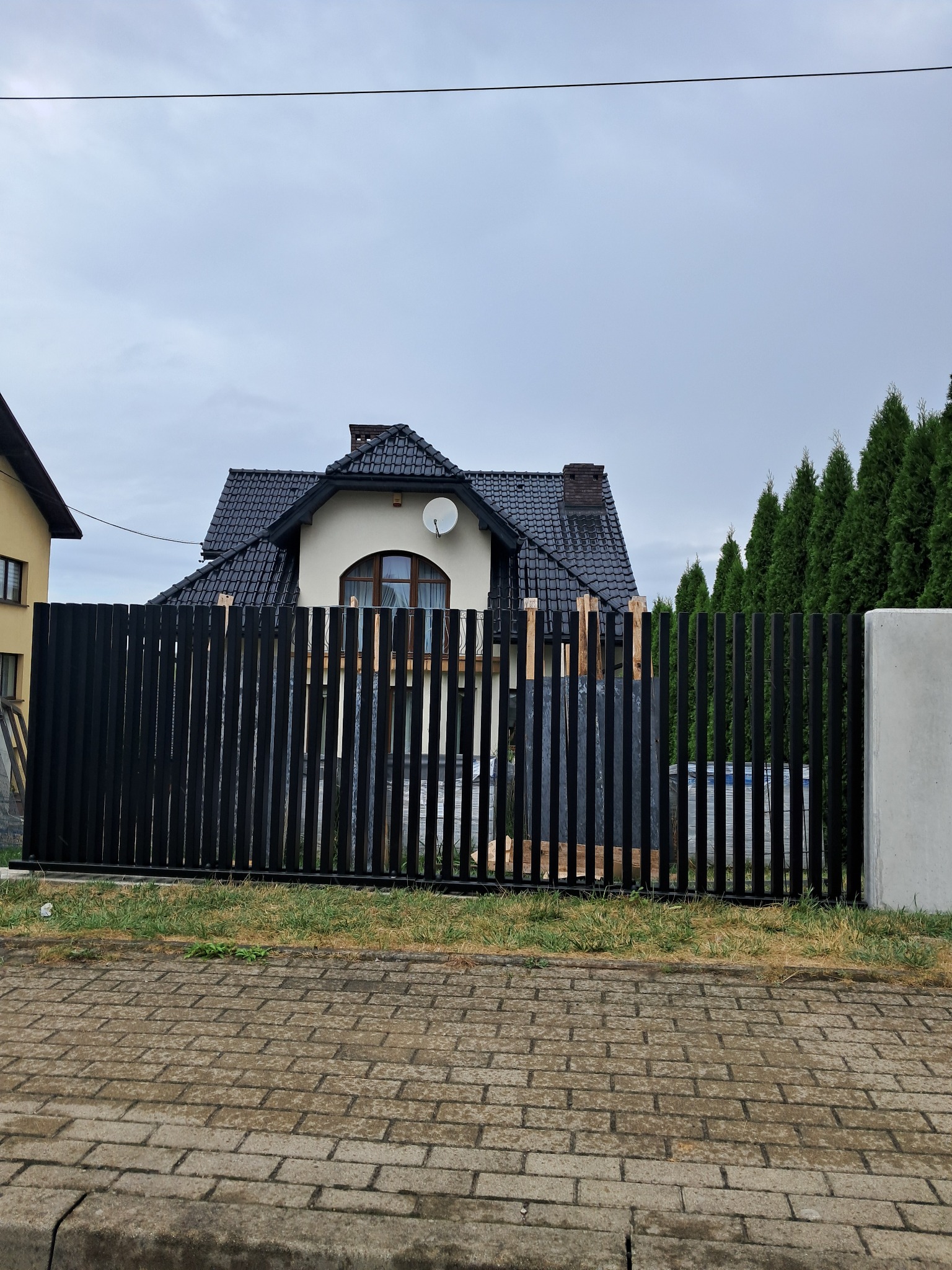 Częściowo zamontowane, czarne, pionowe ogrodzenie metalowe na tle domu z ciemnym dachem i tujami. Widoczne drewniane elementy montażowe.