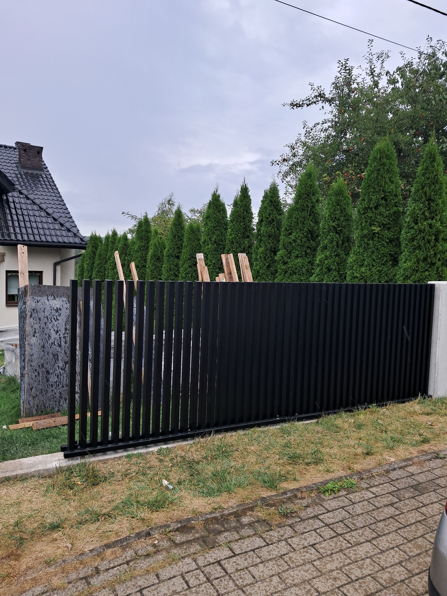 Częściowo zamontowane, czarne, pionowe panele ogrodzenia metalowego na tle zielonych drzew i domu z ciemnym dachem. Widoczne elementy montażowe.